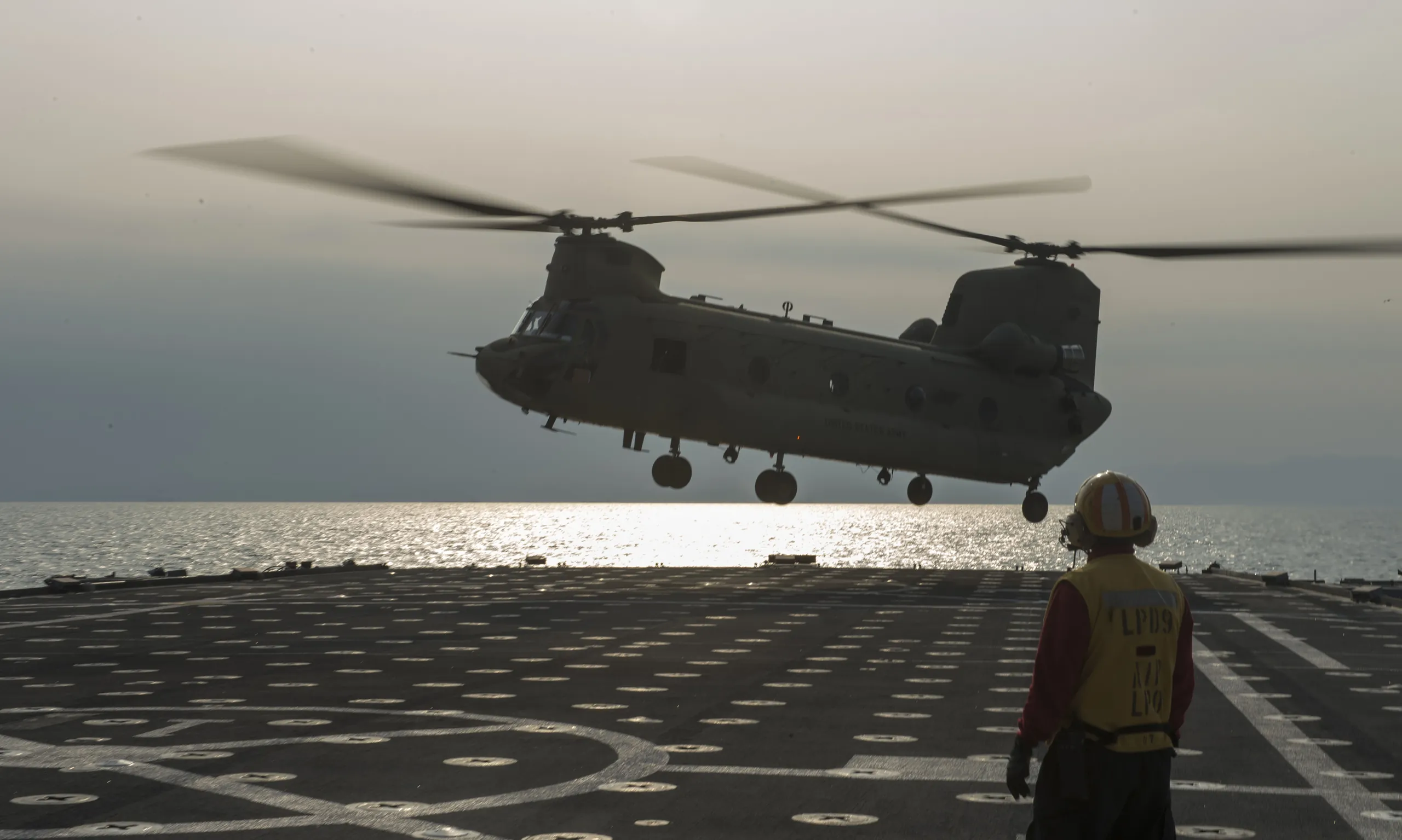 An Army CH-47F lifting off the flight deck of a Navy amphibious ship, a deck crewman in the foreground and the sea behind