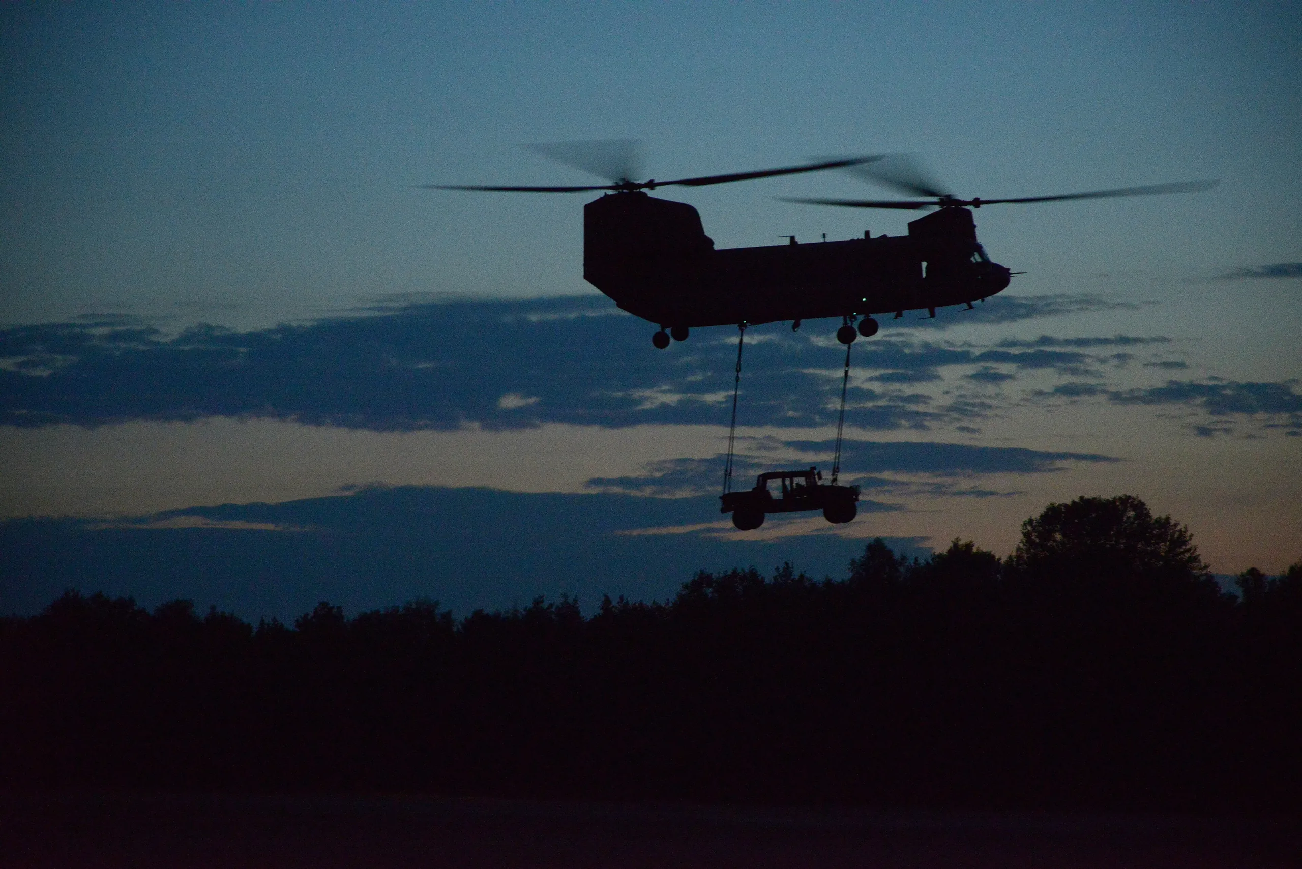 A CH-47F Chinook at dusk carrying a vehicle slung from its cargo hooks, above a dark treeline